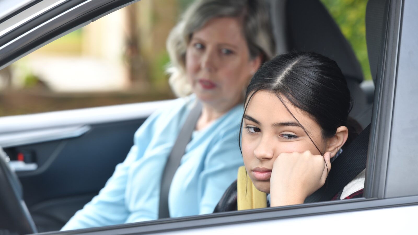 Woman and teenage girl sit in car. Teenage girl annoyed.